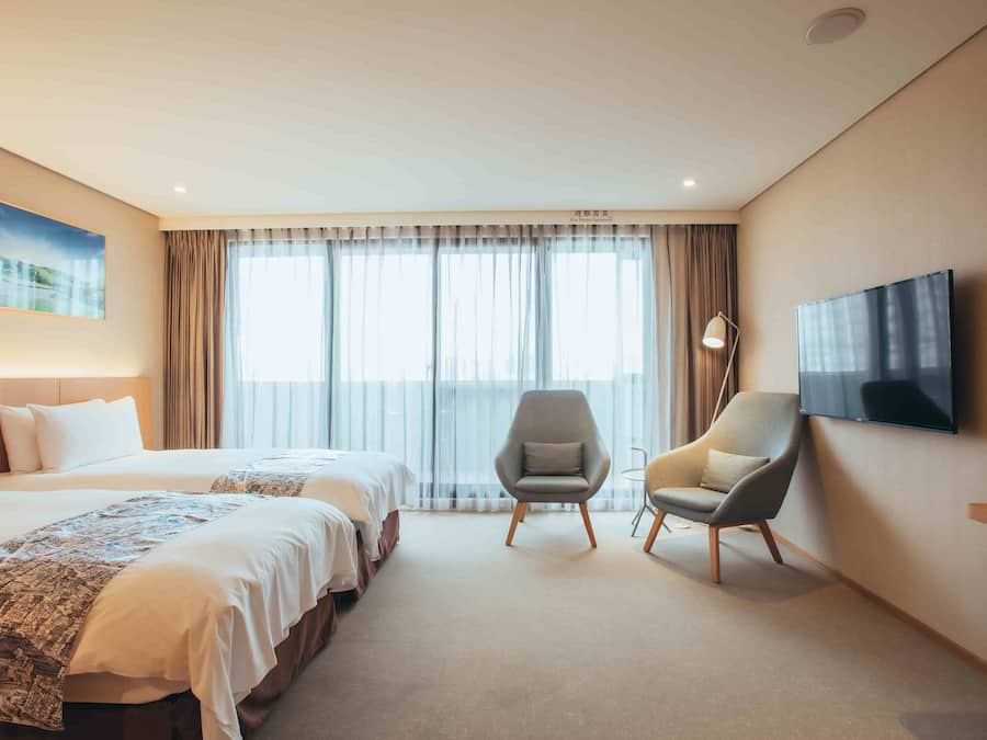 A hotel room with two beds, a flat-screen TV mounted on the wall, and a large window with curtains.