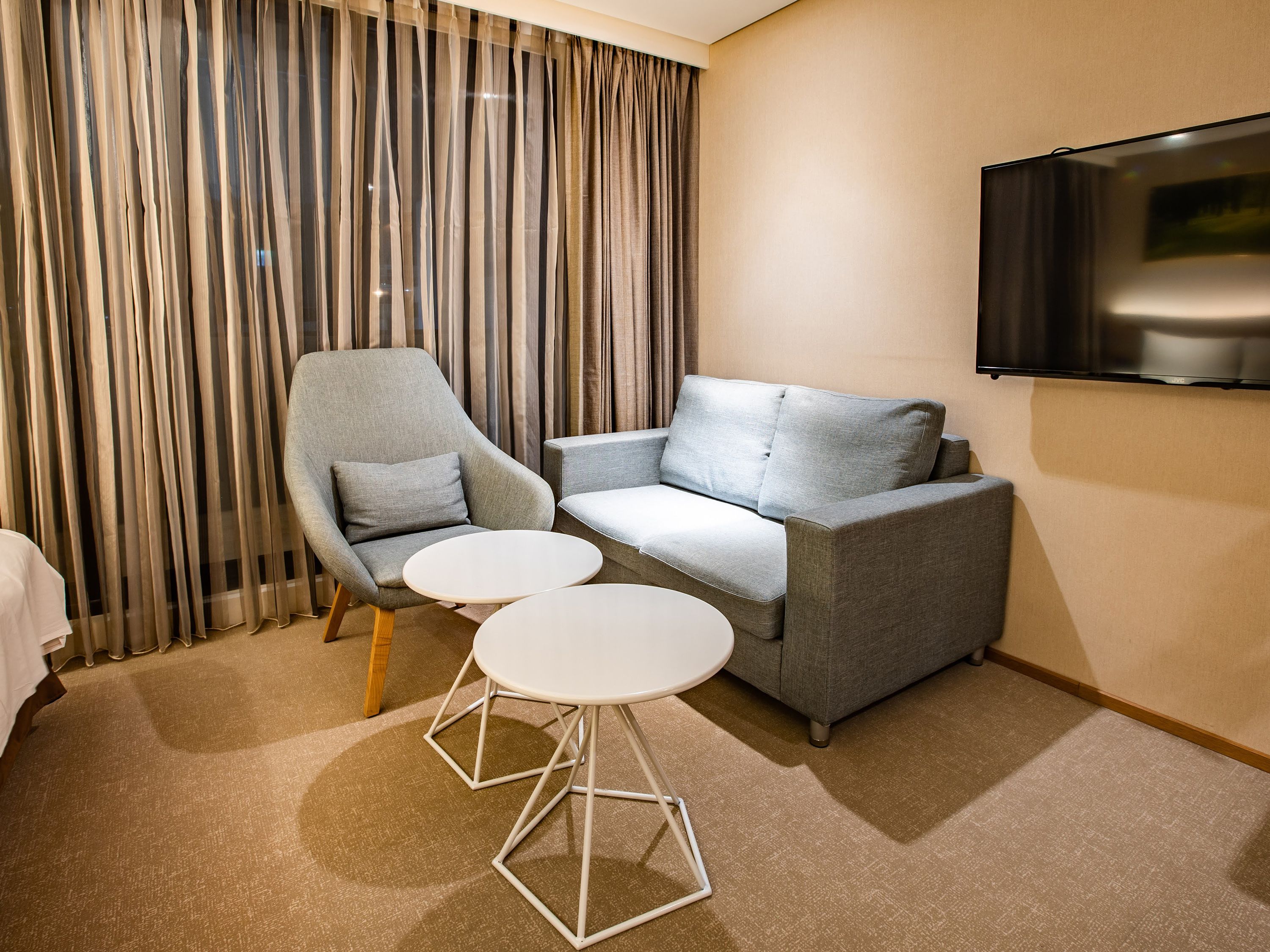 A modern hotel room with a grey sofa, a single armchair, two small round tables, and a flat-screen TV mounted on the wall.