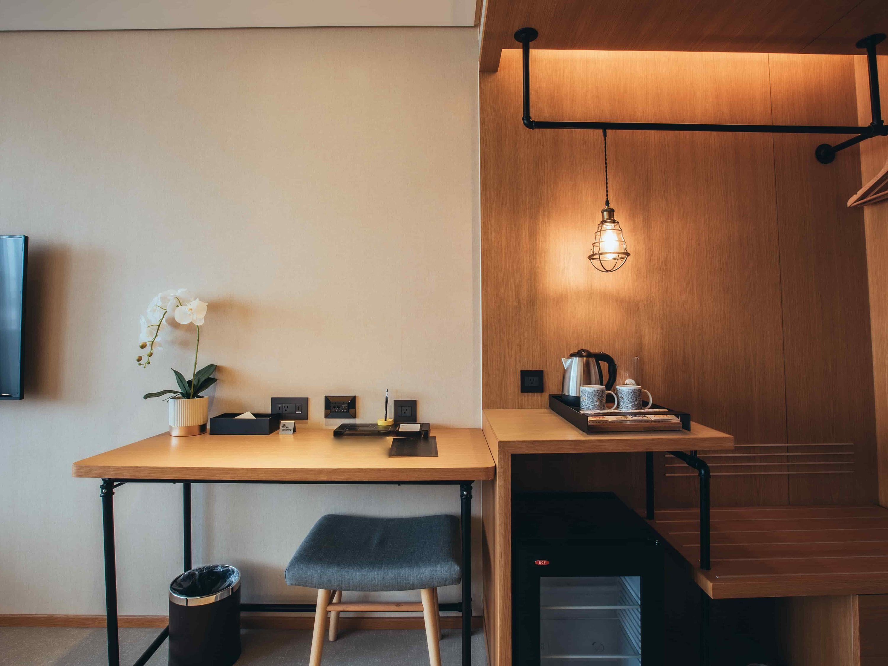 A compact workspace with a wooden table, a single chair, a trash bin, a small potted orchid, a coffee maker, and a hanging light fixture.