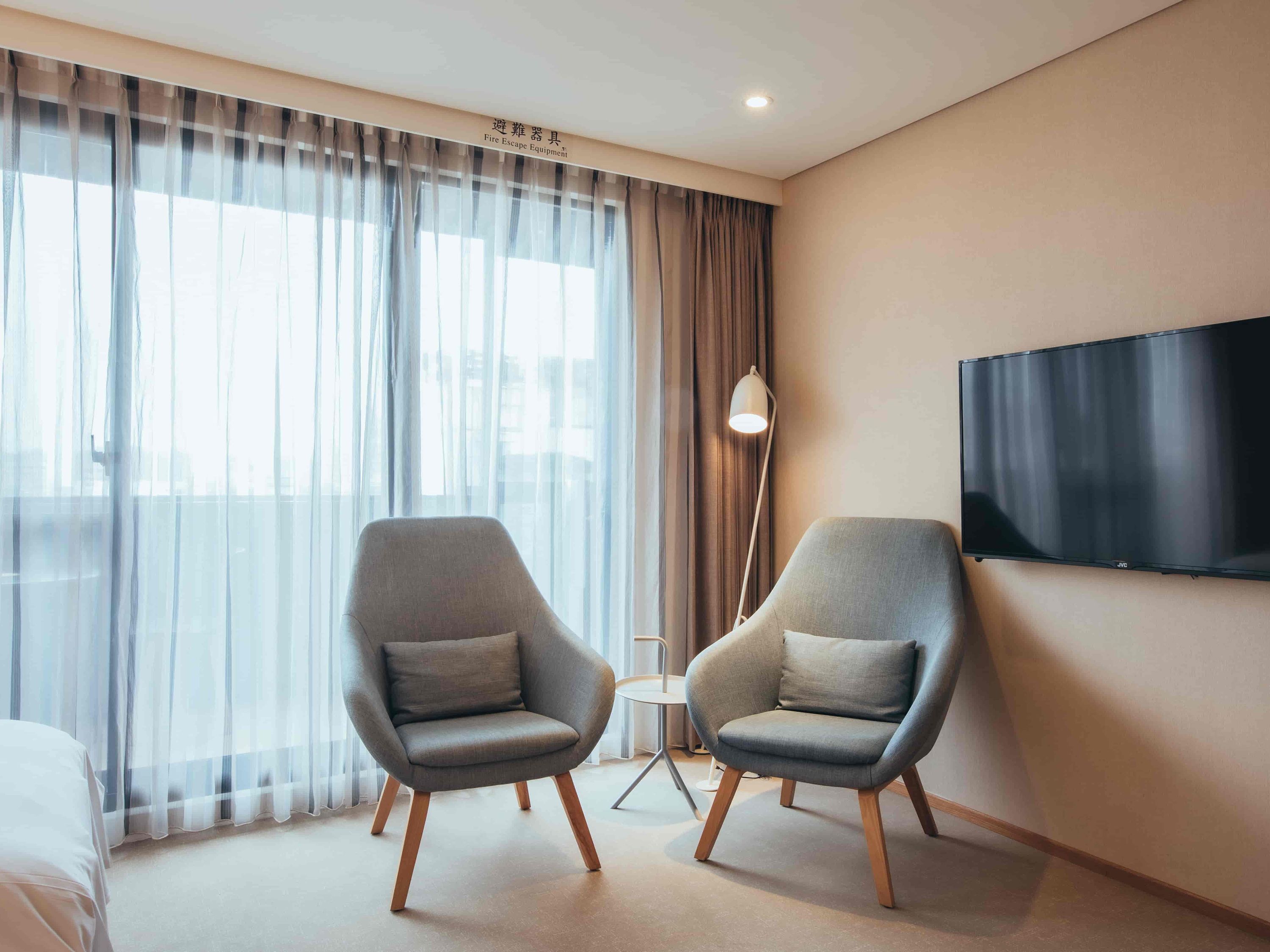 A modern hotel room with two grey armchairs, a flat-screen TV, a floor lamp, and a large window with sheer curtains.