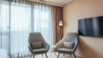 A modern hotel room with two grey armchairs, a flat-screen TV, a floor lamp, and a large window with sheer curtains.