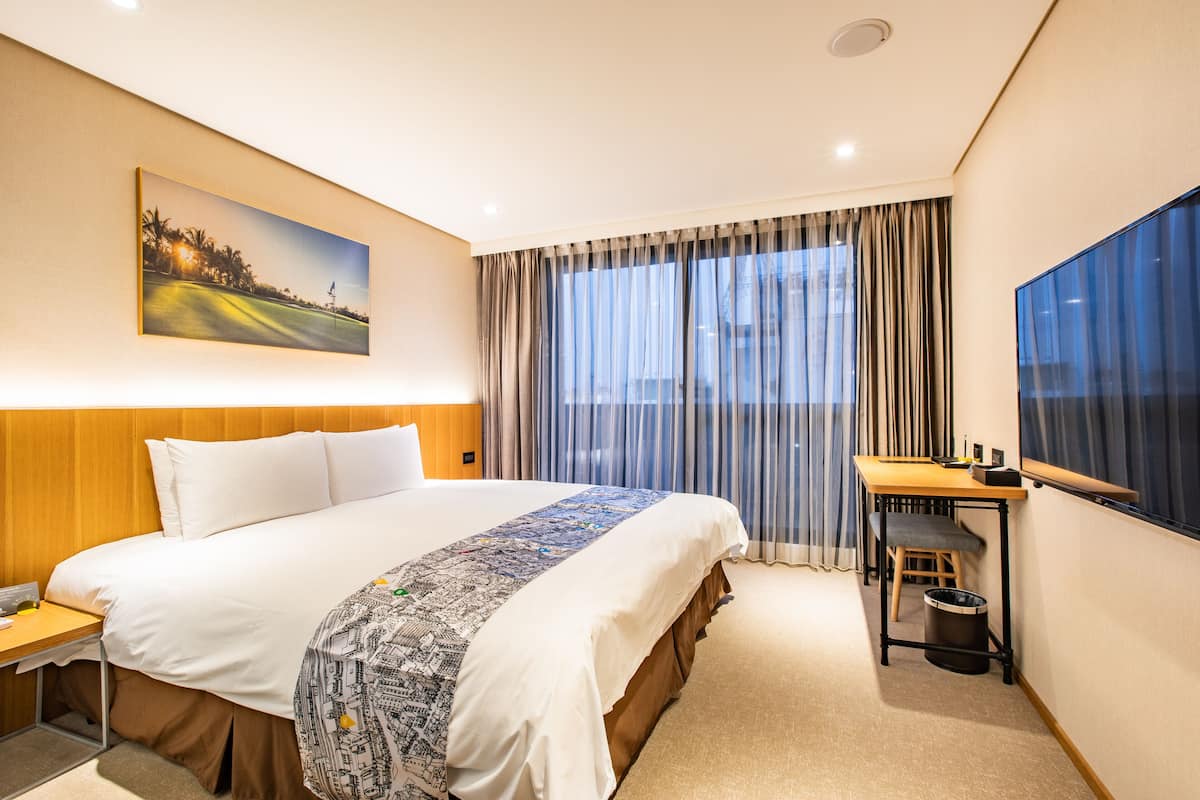 Una habitación de hotel con una cama grande, un escritorio, una silla, un cesto de basura y un cuadro en la pared.