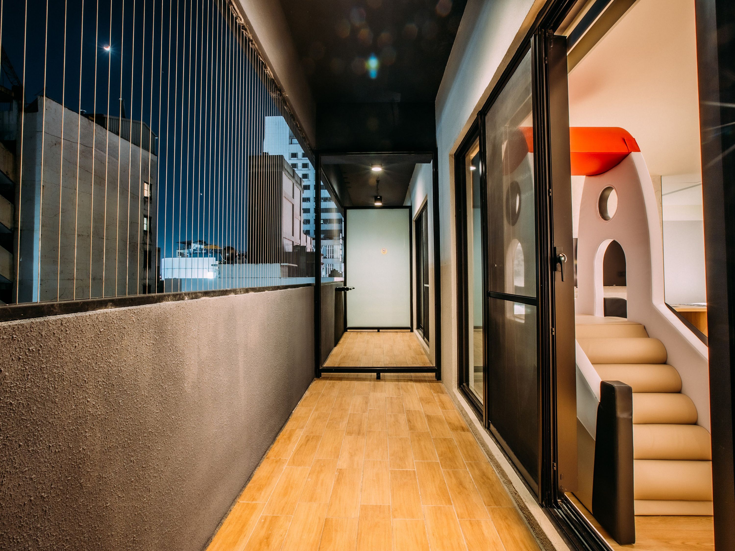 A corridor with a wooden floor, a glass door leading to a room with a staircase, and a view of the outside through a window with vertical blinds.