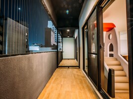 Un pasillo con piso de madera, una puerta de vidrio que da a una habitación con una escalera, y vistas al exterior a través de una ventana con persianas verticales.
