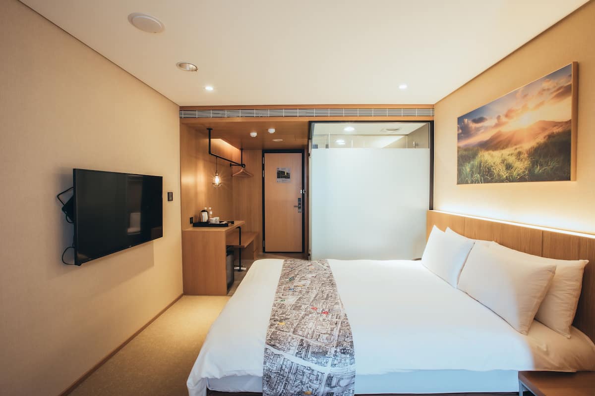 Una habitación de hotel moderna con una cama grande, un televisor de pantalla plana, un escritorio de madera y un baño visible a través de una puerta abierta.