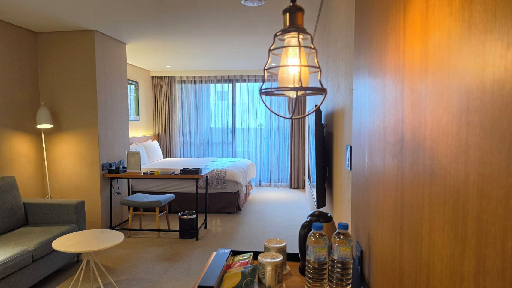 A hotel room with a bed, a desk, a chair, a sofa, a table, a lamp, and a TV.