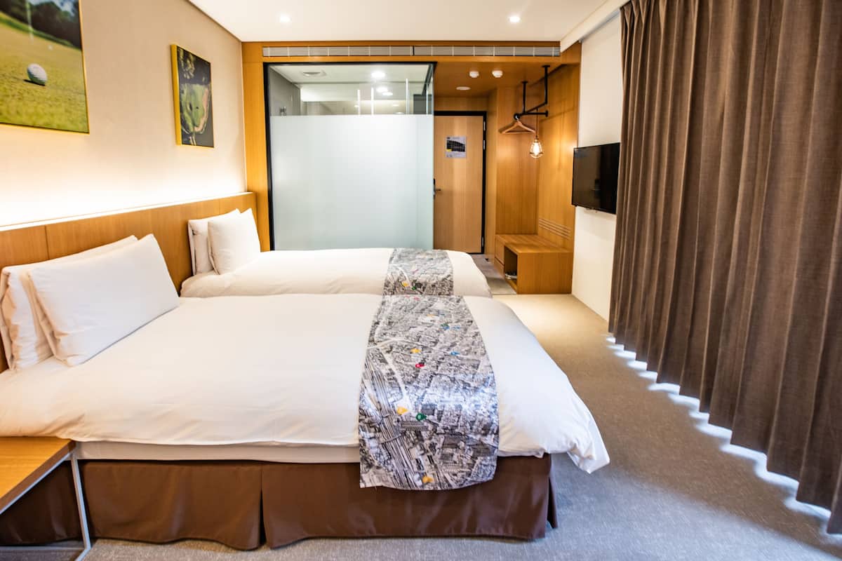 Habitación de hotel con dos camas, un ventanal grande, un televisor y un banco de madera.