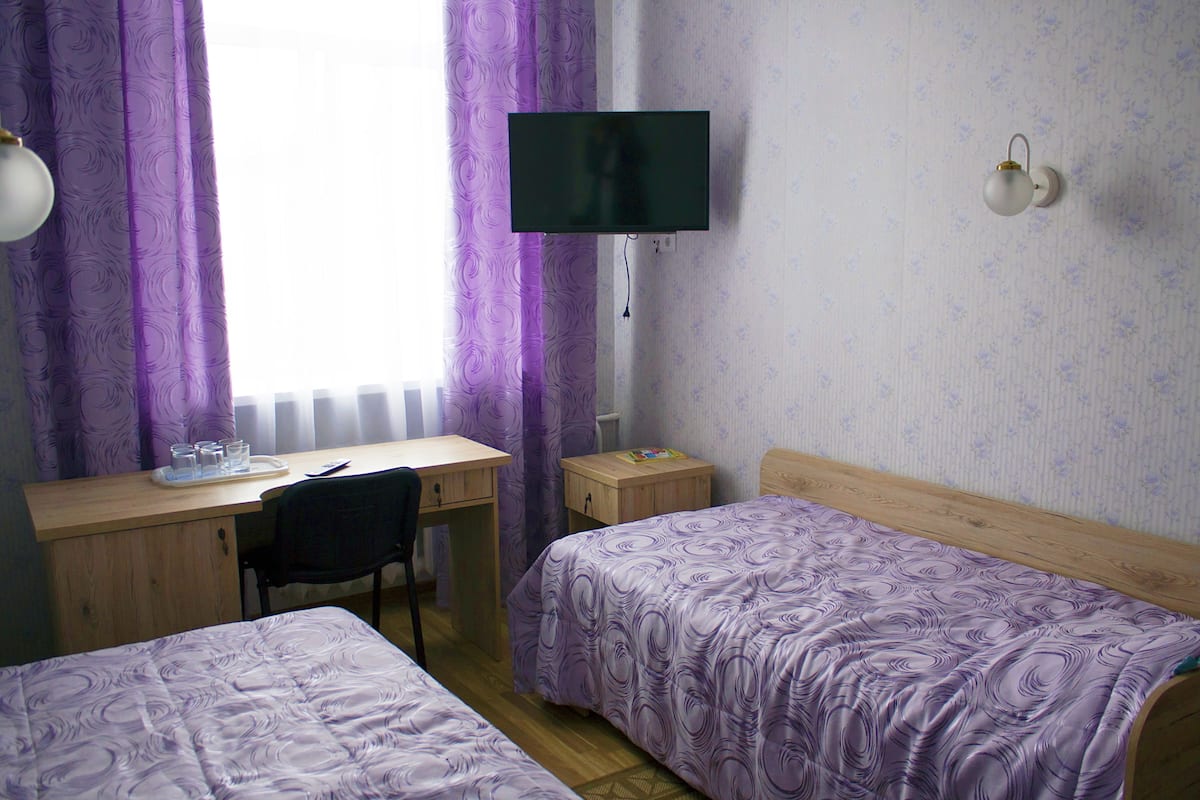 Una habitación con dos camas, un escritorio, una silla, un televisor instalado en la pared y una ventana con cortinas moradas.