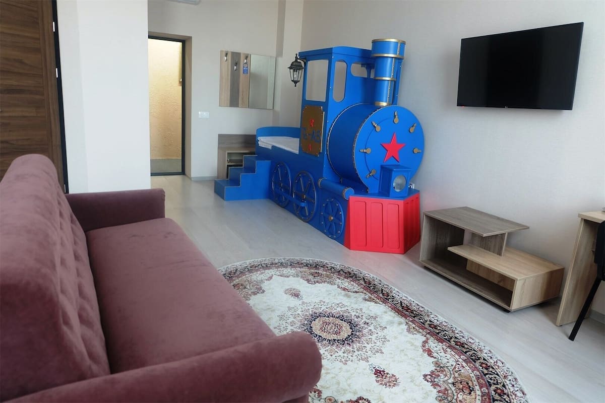 Una sala de estar con un modelo de tren azul, un televisor de pantalla plana, un sofá rojo y una alfombra estampada.