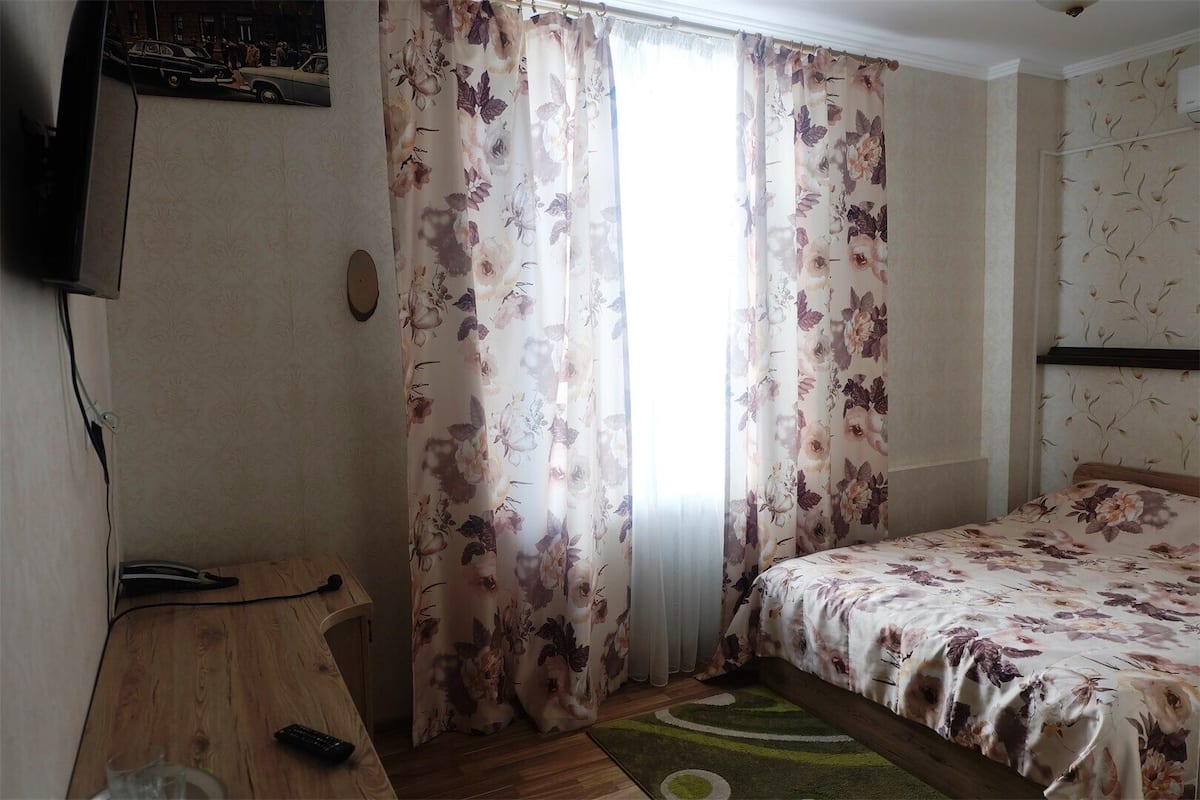 Un dormitorio con cama, una mesita de noche de madera, televisor montado en la pared y cortinas con motivos florales.