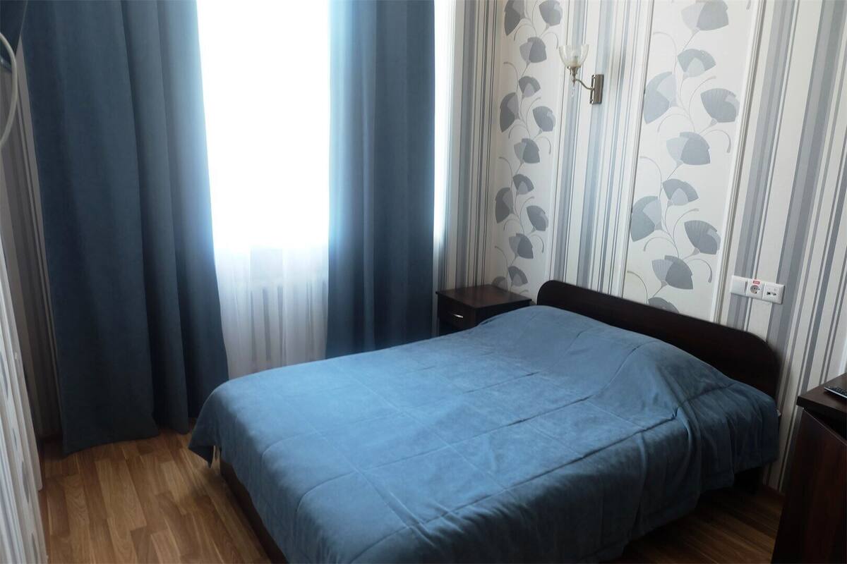 Un dormitorio con cama, piso de madera, cortinas, una mesita de noche y un muro con estampado de hojas.
