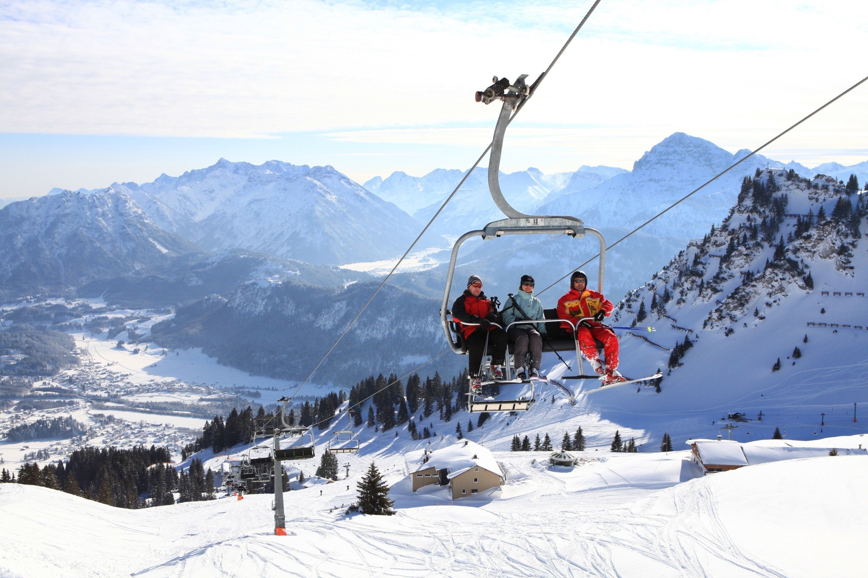 Eine Sesselbahn mit Fahrgästen, schneebedeckte Berge und eine kleine Hütte im Hintergrund.