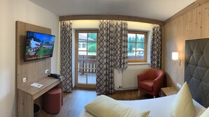 A hotel room with a wooden headboard, a desk with a chair, a television, and a window with patterned curtains. - Gästehaus am Graben (Hoefen)