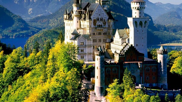 A castle with multiple towers, surrounded by lush greenery and mountains in the background. - Gästehaus am Graben (Hoefen)