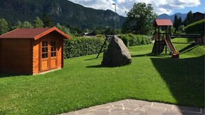 A wooden cabin, a playground with a slide, and a stone pathway in a mountainous area. - Gästehaus am Graben (Hoefen)