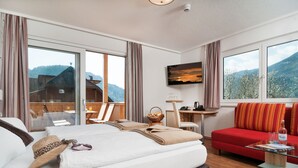 A hotel room with a bed, a red sofa, a desk, and a large window offering a mountain view. - Wellness Landhaus PARTH (Ossiach)