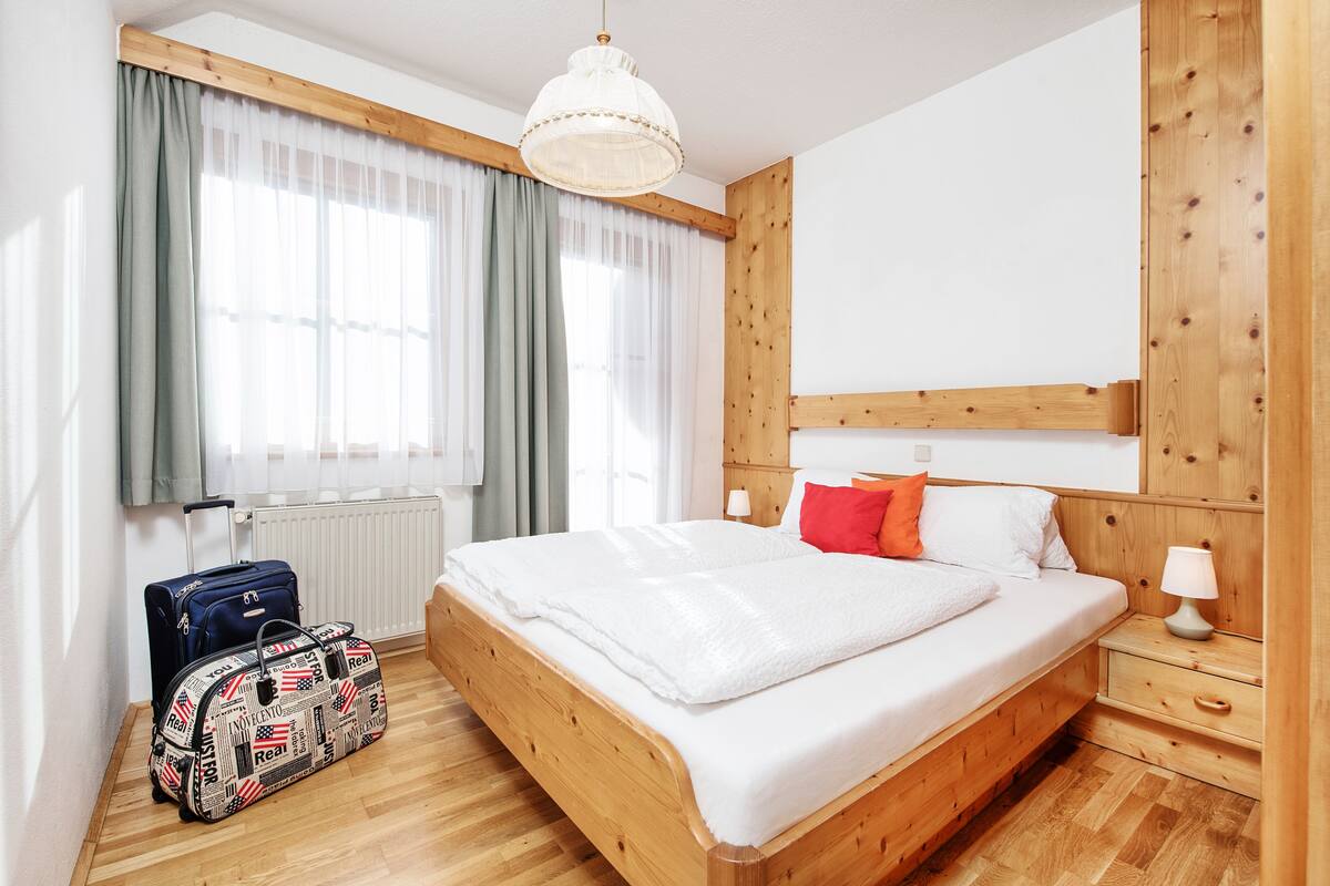 Un dormitorio con una cama de madera, una mesita de noche, una lámpara, una maleta y una ventana con cortinas.