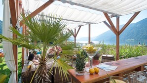 A covered outdoor seating area with a table, chairs, and a view of a lake and mountains. - Wellness Landhaus PARTH (Ossiach)