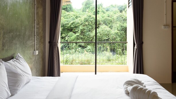 Habitación de hotel con una cama grande, ropa de cama blanca, vista a espacios verdes a través de una puerta balcón y un equipo de aire acondicionado sobre la puerta.