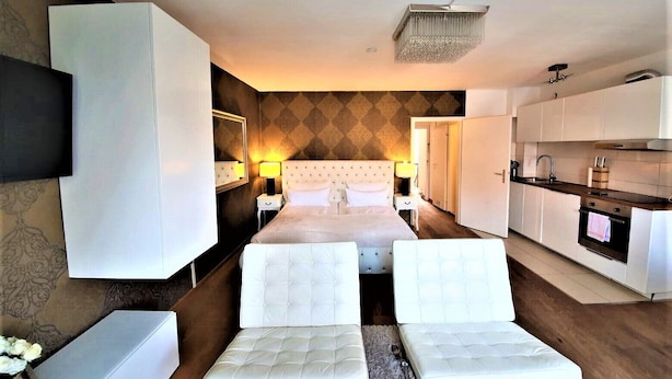Una habitación de hotel moderna con una cama grande, una zona de estar con dos sillones blancos acolchados, una pequeña cocina con estufa y fregadero, y un televisor montado en la pared.