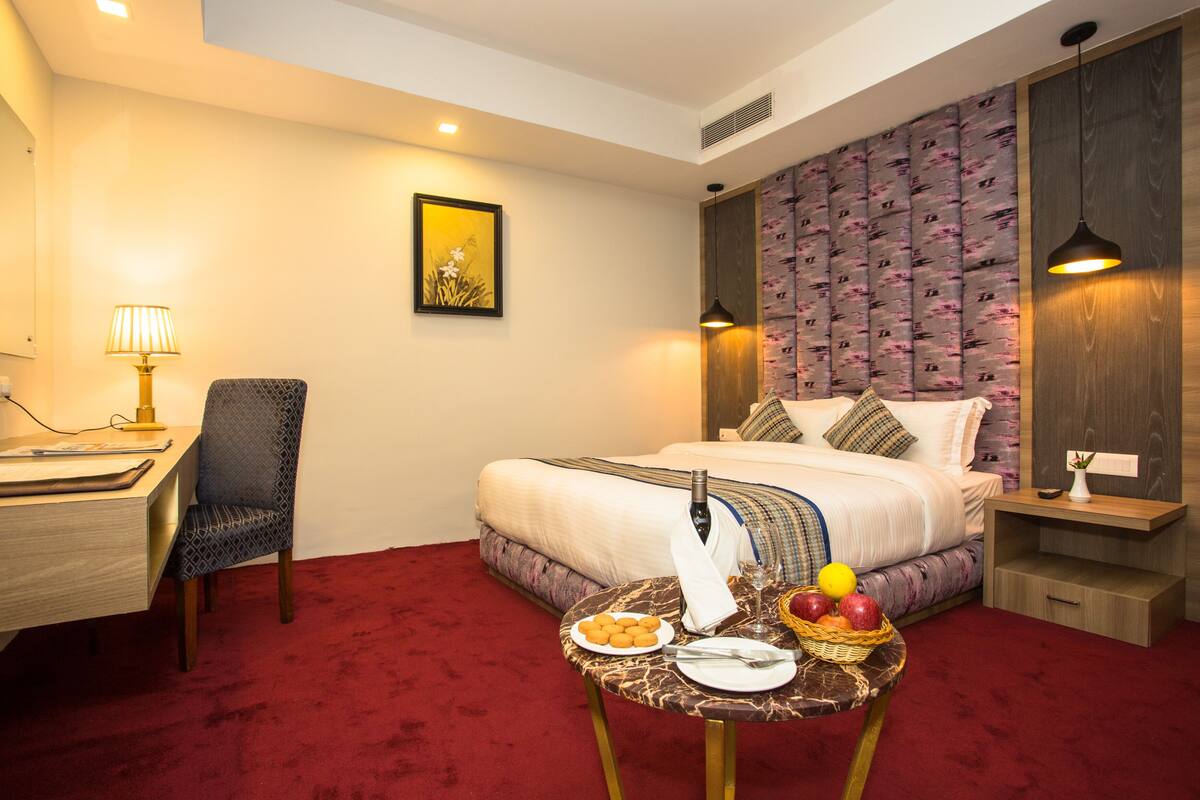 A hotel room with a bed, a desk, a chair, a small table with food and drinks, and a painting on the wall.