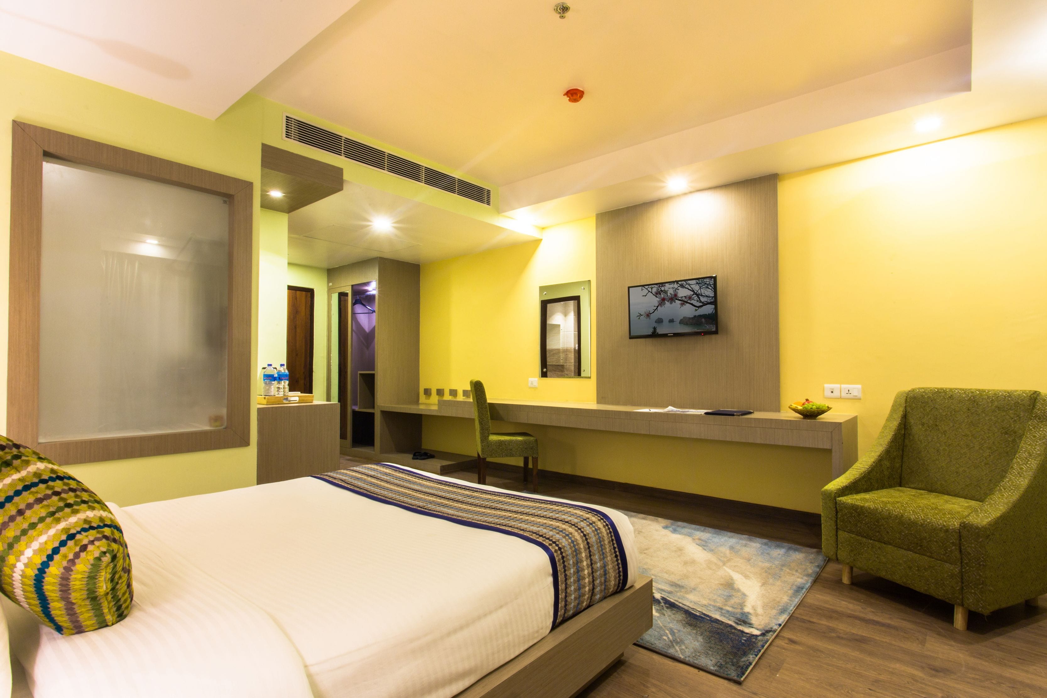 Une chambre d’hôtel avec un lit, un fauteuil vert, un bureau avec une chaise, une télévision et une salle de bain visible à travers une porte ouverte.