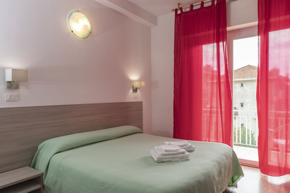 Ein ordentlich bezogenes Bett mit einer grünen Tagesdecke und weißen Handtüchern, eine Wandleuchte sowie ein Fenster mit roten Vorhängen.