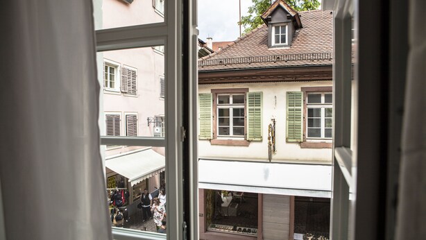A view through a window of a building with a shop on the ground floor and a balcony with a railing. - New England Apartment (Freiburg im Breisgau)