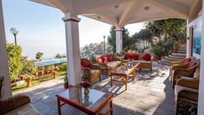 A covered outdoor seating area with wicker furniture and a glass table, overlooking a scenic landscape. - Mirabel Resort (Dhulikhel)