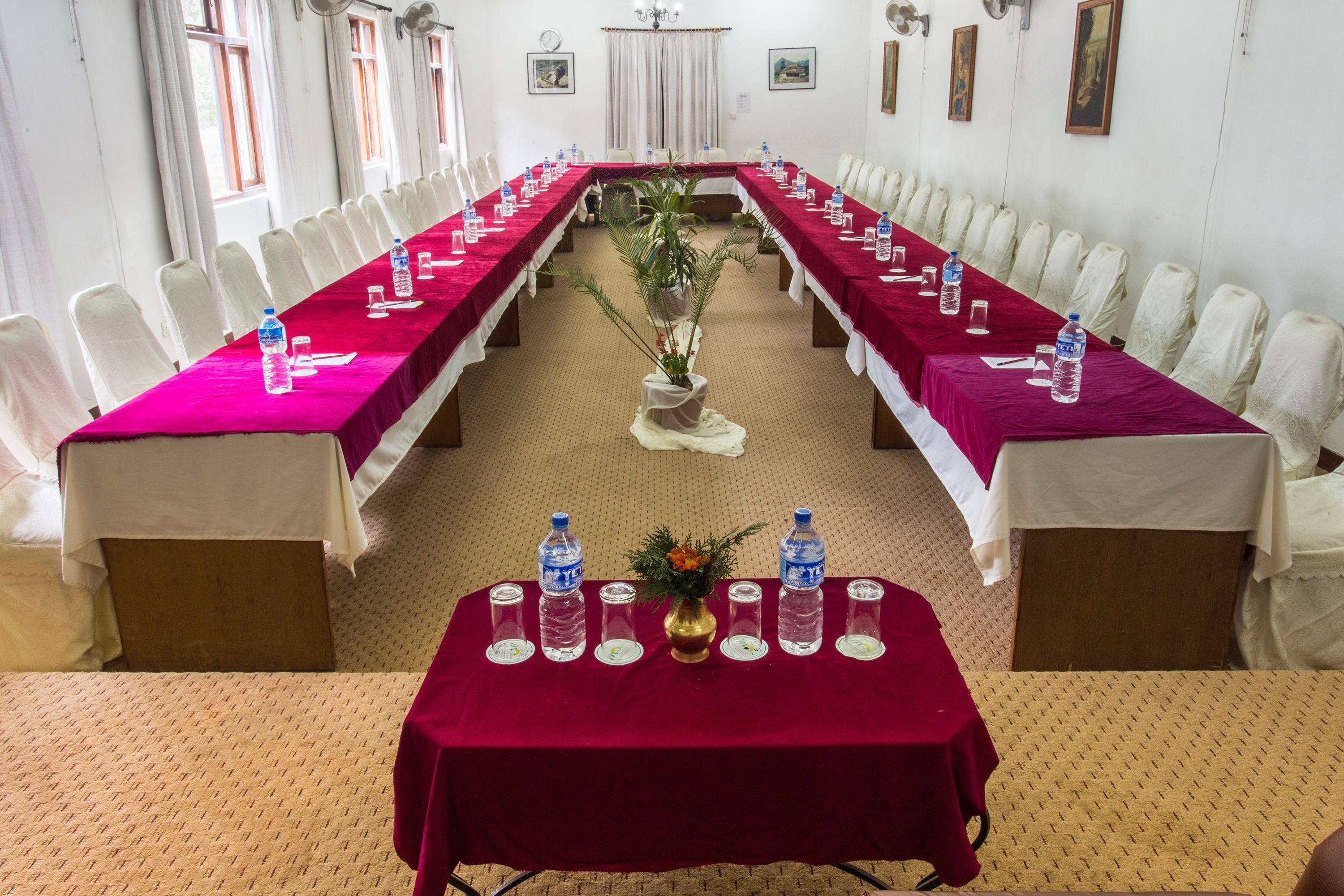 Une salle de conférence avec de longues tables, des chaises, des bouteilles d’eau et un vase rempli de fleurs.