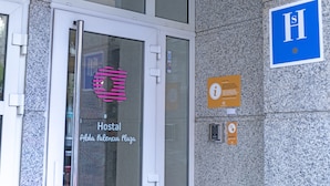 Rakennuksen sisäänkäynti, jossa on lasiovi, jonka yläpuolella on kyltti, jossa lukee ”Hostal Alda Palacio Plaza”. Sen vieressä on sininen kyltti, jossa on kirjain ”H”, mikä viittaa sairaalaan, sekä pieni tiedotustaulu.