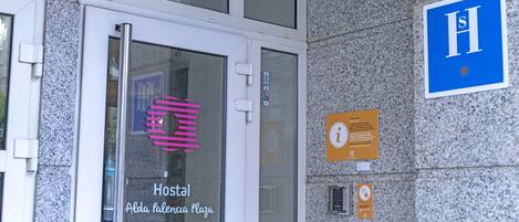 A building entrance with a glass door labeled "Hostal Alda Palacio Plaza," a blue sign with "H" indicating a hospital, and a small information board.