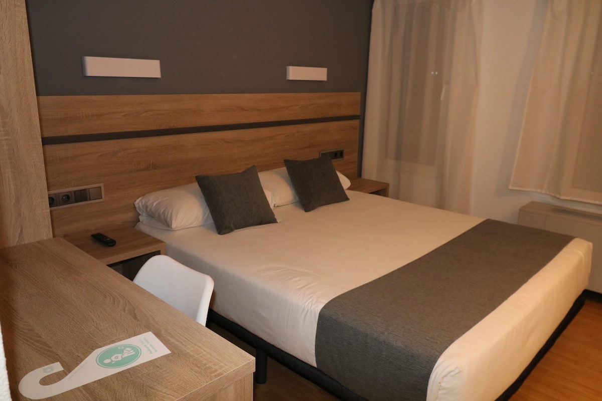 Ein Hotelzimmer mit einem hölzernen Kopfteil, einem großen Bett mit grau-weißer Bettwäsche, einem Nachttisch mit Telefon und einem kleinen Schreibtisch mit Stuhl.