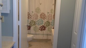 Un couloir étroit mène à une salle de bain équipée de toilettes, d’un rideau de douche et d’un tapis à rayures.