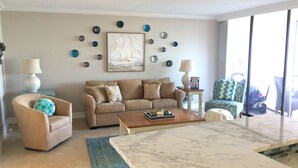 Ein Wohnzimmer mit einem beigen Sofa, einem Couchtisch und einem Teppich mit Muster. An der Wand befinden sich dekorative Teller und auf einem Beistelltisch steht eine Lampe.