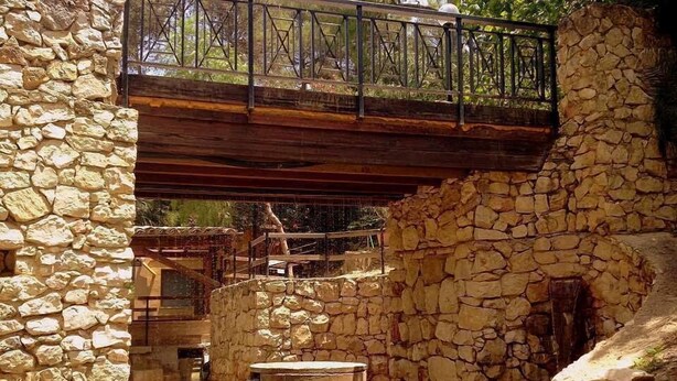 Un pati de pared de pedra amb un pont de fusta, una petita font i una banca.