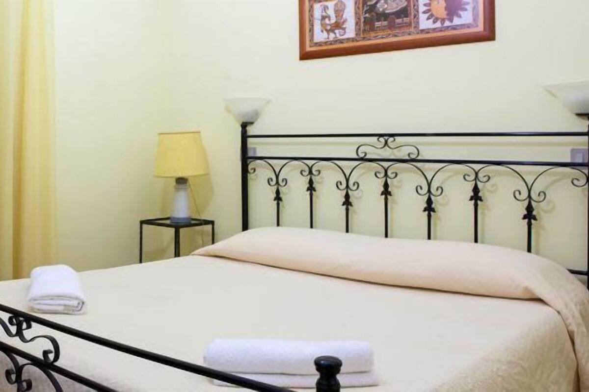 A bed with a metal frame and a beige bedspread, a small bedside table with a lamp, and a chair to the left.