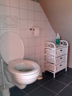Un baño con inodoro blanco, dispensador de papel higiénico blanco montado en la pared y un mueble de almacenamiento de mimbre blanco con una botella encima.