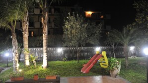 Un espacio exterior nocturno con un parque infantil, que incluye un tobogán y columpios, rodeado de palmeras y plantas en macetas.