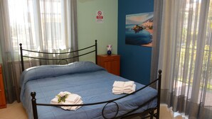 Una cama bien hecha con ropa de cama azul y toallas blancas, una mesita de noche de madera con un jarrón, y un cuadro de una escena costera en la pared.