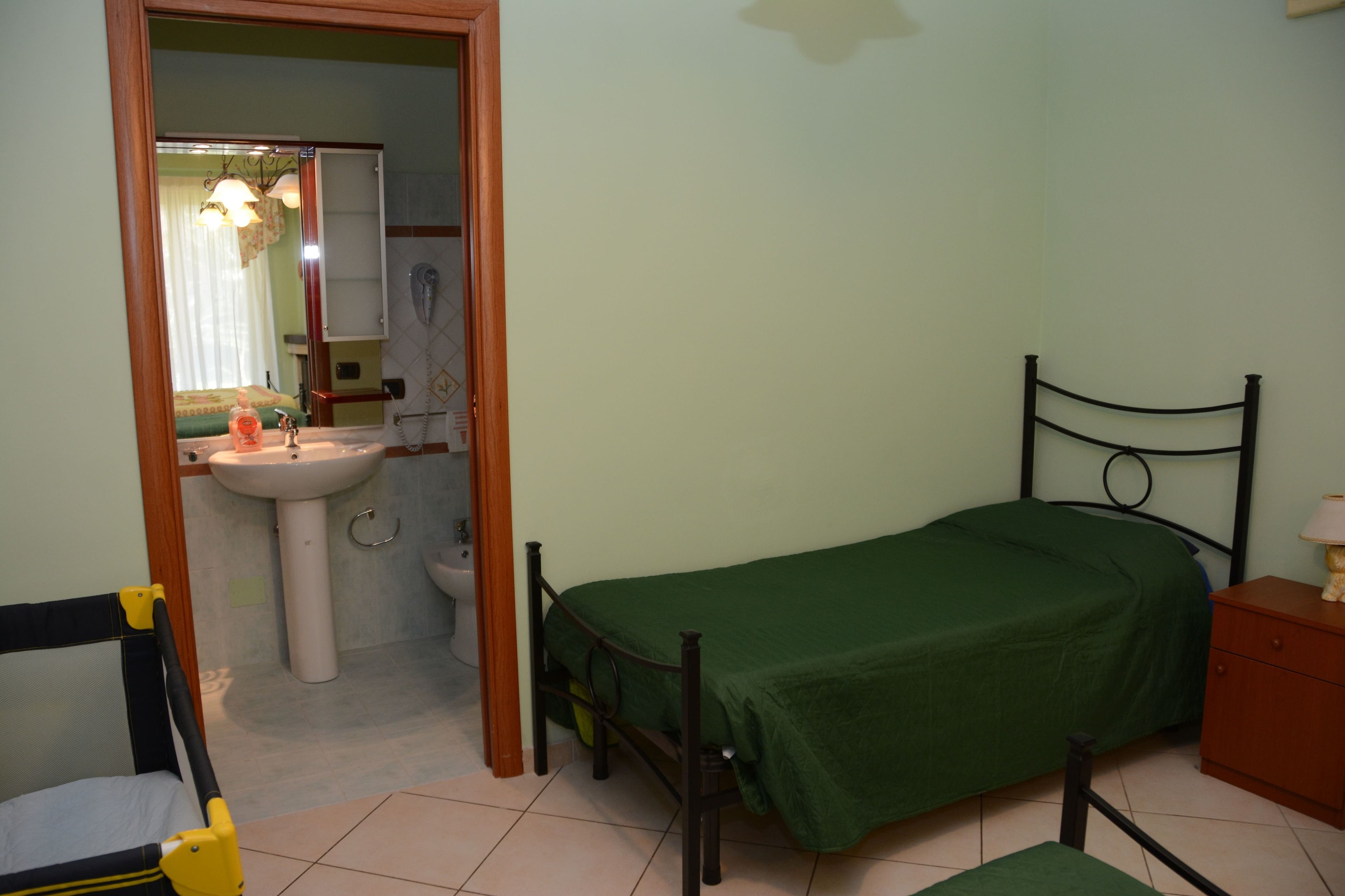 Una camera piccola con un letto singolo, un comodino, una sedia di legno, un bagno con lavandino e water, e uno specchio.