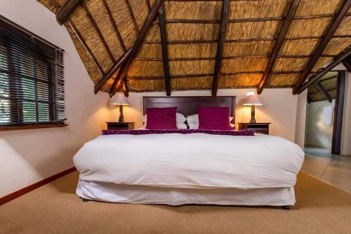 Un dormitorio con techo de paja, una cama grande con ropa de cama blanca y morada, dos mesitas de noche con lámparas y una ventana con persianas.