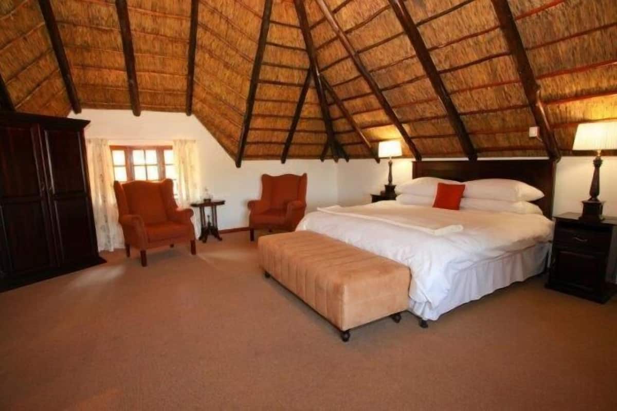 Una habitación con un alto techo de paja, una cama grande con sábanas blancas, dos sillones, una mesita y una mesita de noche con una lámpara.