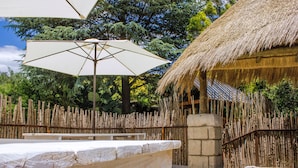 A thatched-roof hut with a bamboo fence, a stone bench, and a large white umbrella. - Rainforest Boutique Camp (Krugersdorp)