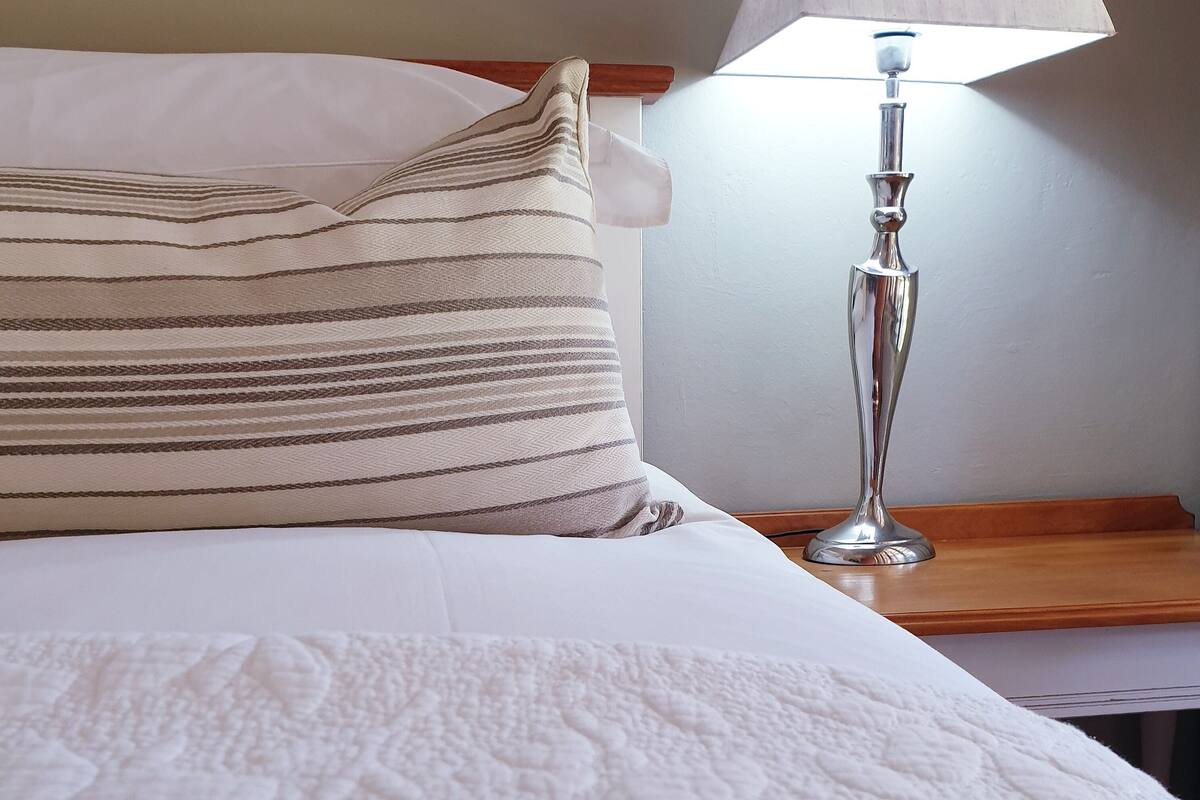 Cama bem arrumada com colcha branca, travesseiro listrado e abajur sobre um criado-mudo de madeira.