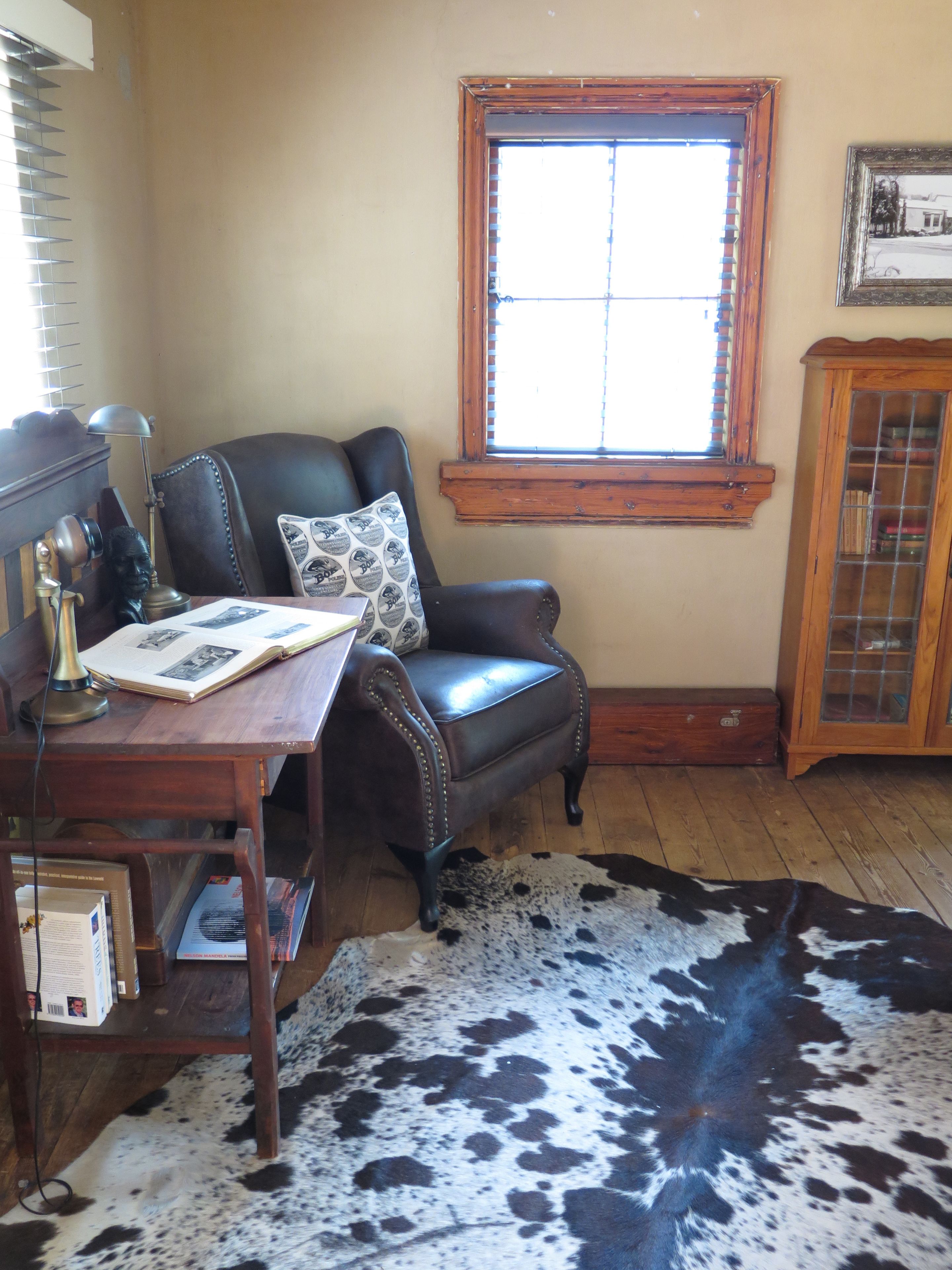 A room with a leather armchair, a wooden desk with a lamp, a cowhide rug, a window with blinds, and a wooden cabinet.