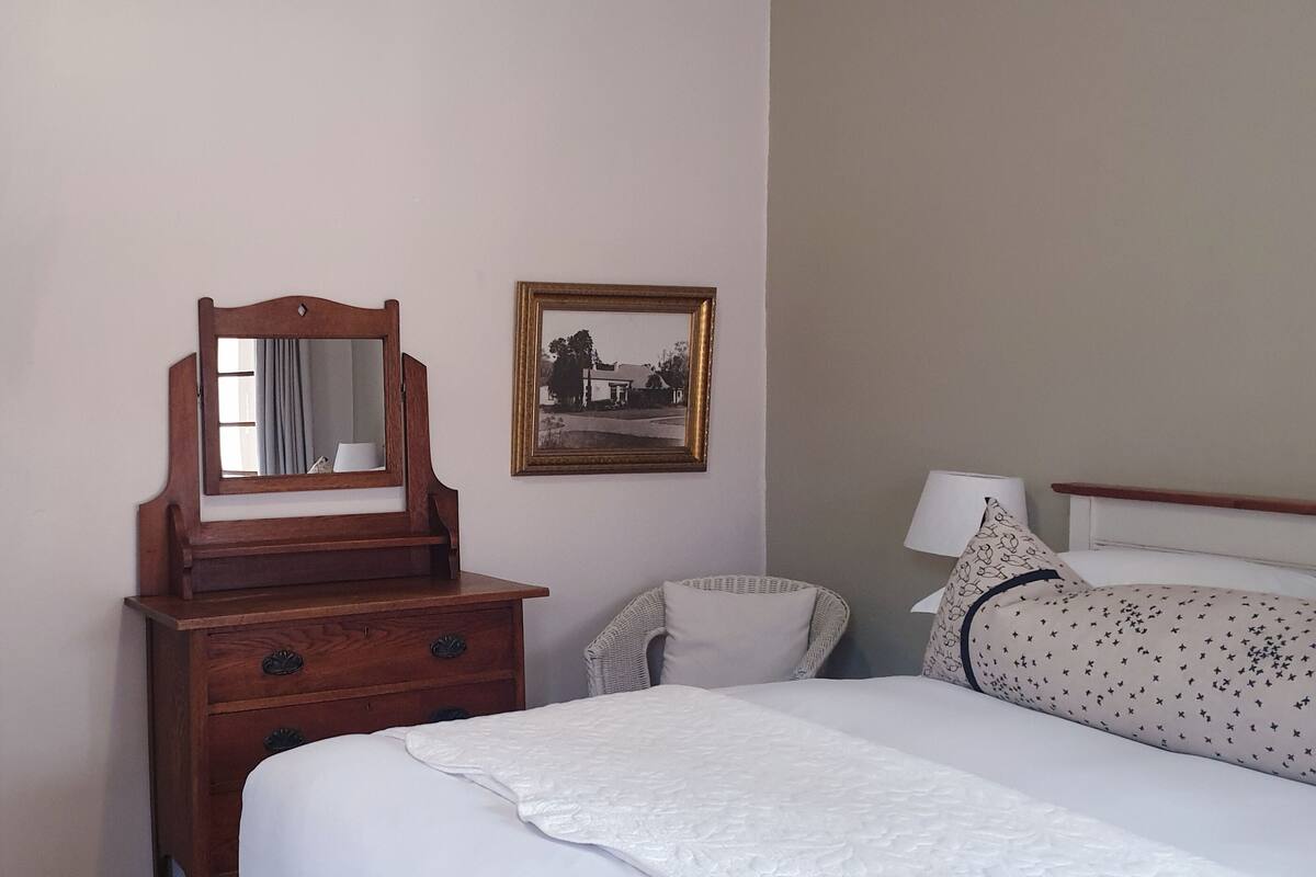 Quarto com uma cama, penteadeira de madeira com espelho, uma cadeira, escrivaninha com chaleira e quadro na parede.