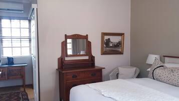 A bedroom with a bed, a wooden dresser with a mirror, a chair, a desk with a kettle, and a framed picture on the wall.
