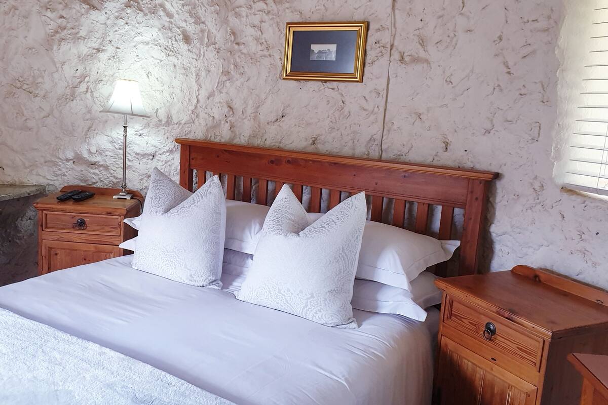 Quarto com cama de madeira, roupa de cama branca, mesa de cabeceira com abajur, espelho e um quadro na parede.