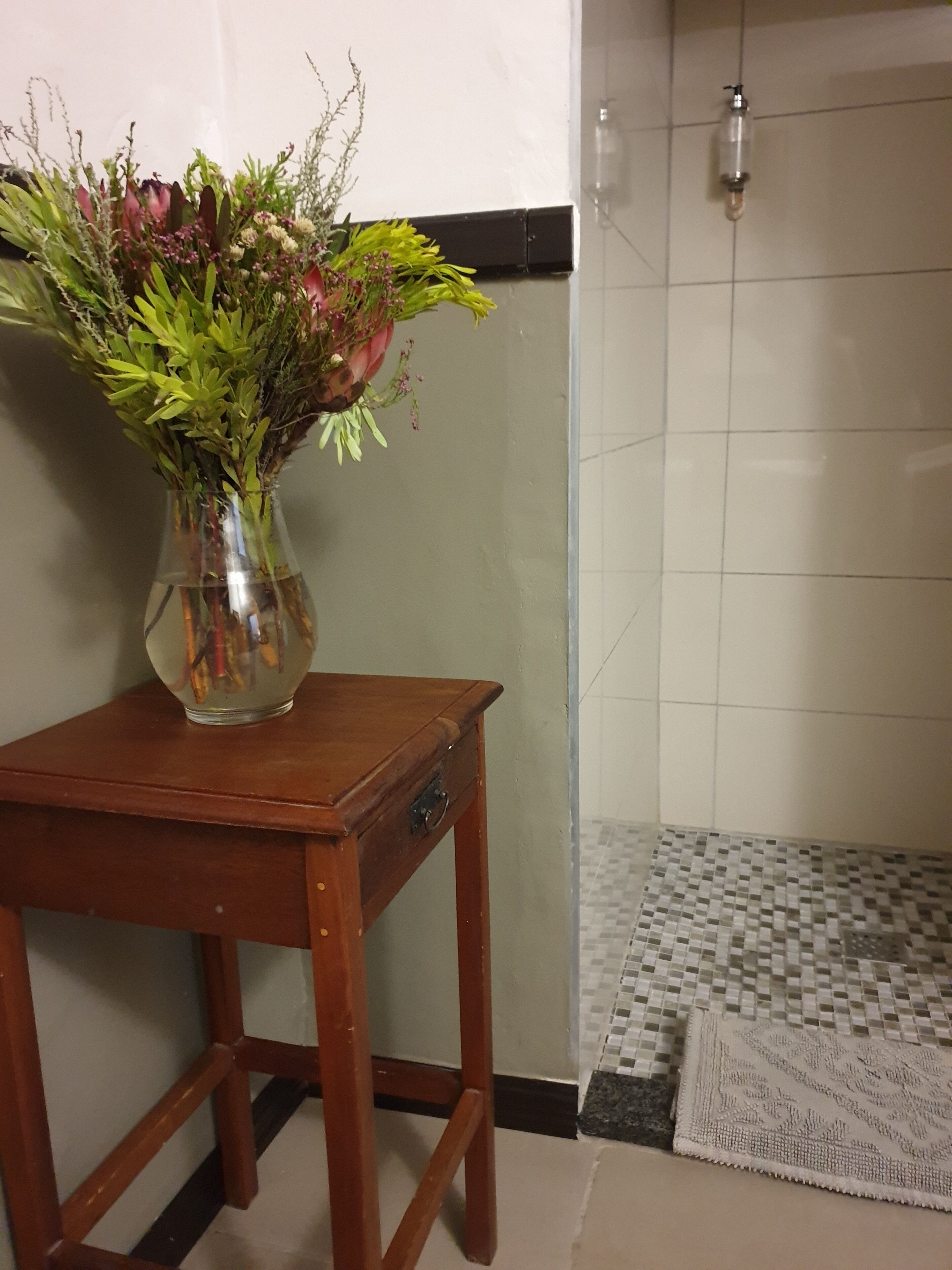 A bathroom with a tiled floor, a shower area, and a wooden table with a vase of flowers.